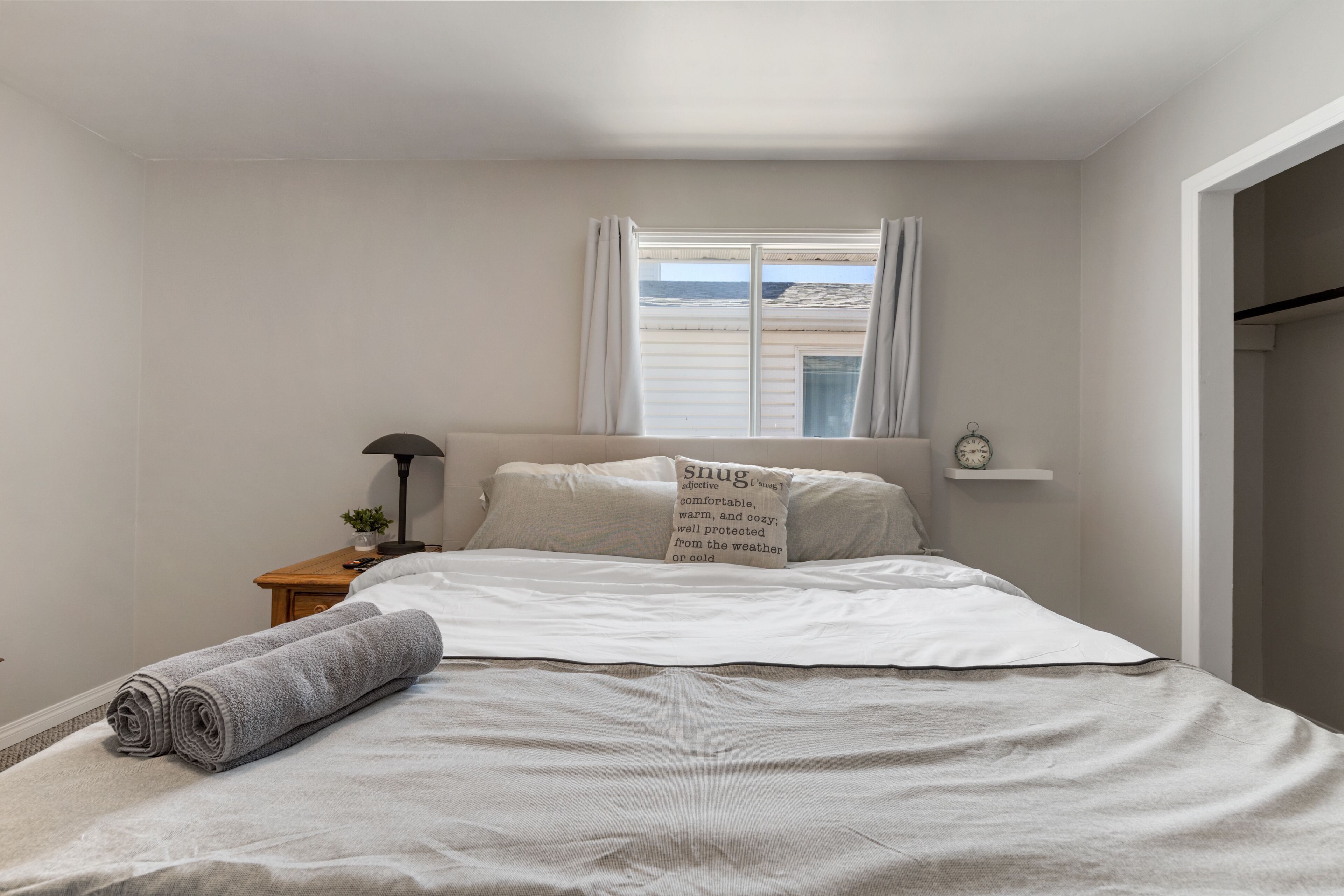 A bedroom with a bed, a nightstand, a lamp, a window with curtains, and a view of the sea.