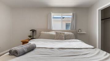 A bedroom with a bed, a nightstand, a lamp, a window with curtains, and a view of the sea.