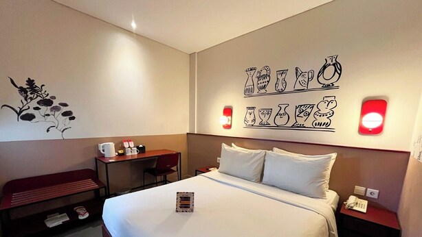 A hotel room with a large bed, a desk, a chair, and a bench. There are wall decorations and a nightstand with a lamp. - Kotta GO Yogyakarta (Yogyakarta)
