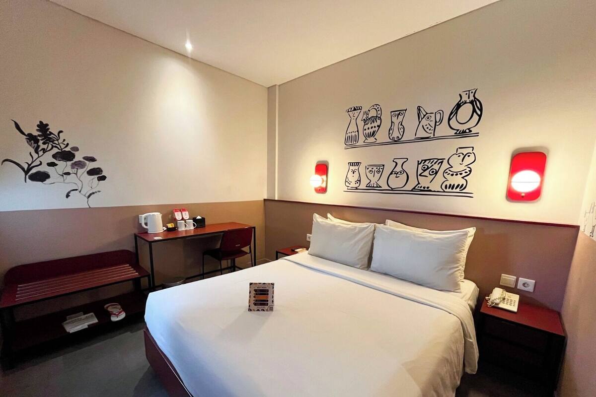 Ein Hotelzimmer mit einem großen Bett, einem Schreibtisch, einem Stuhl und einer Bank. An den Wänden befinden sich Dekorationen und ein Nachttisch mit einer Lampe.
