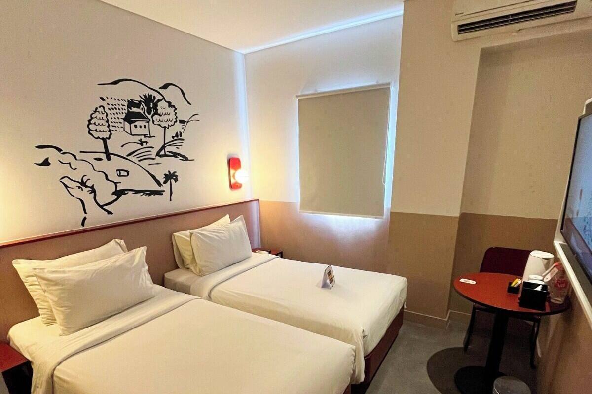 Ein Hotelzimmer mit zwei Betten, einem kleinen Tisch, einem Mülleimer, einer Wand-Klimaanlage und einer Wanddekoration.