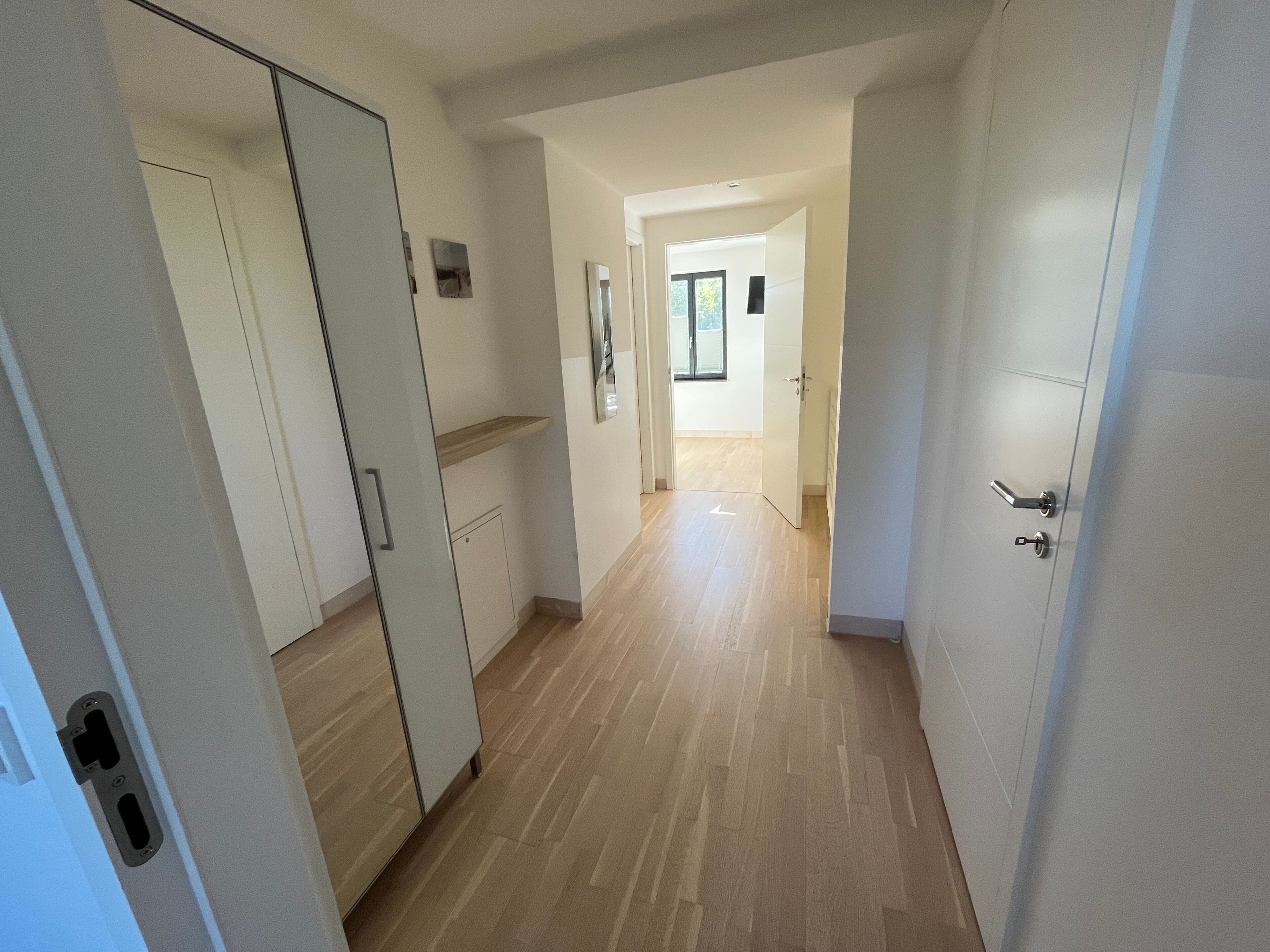 A corridor with a wardrobe, a door, and a hallway leading to a room with a window.