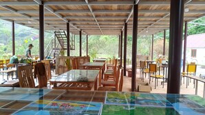 An outdoor dining area with wooden tables and chairs, a glass-top table with a map, and a view of greenery and a building in the background.