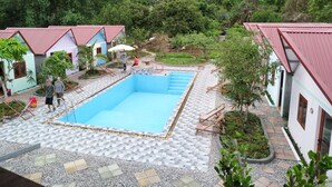 A pool area with tiled flooring, surrounded by small buildings with red roofs, and lush greenery in the background.