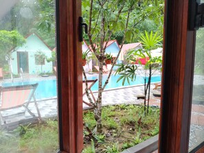 A view through a window with a wooden frame, looking out to a swimming pool area with a tiled deck, a small tree, and some potted plants. - Occult Lan Ha Farmstay Mt (Hai Phong)