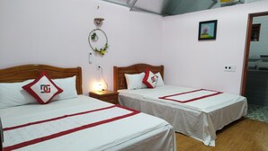 A hotel room with two beds, each with a wooden headboard, white bedding, and red accents. There is a small table with a lamp between the beds, and a wall-mounted light fixture above the bed on the left.