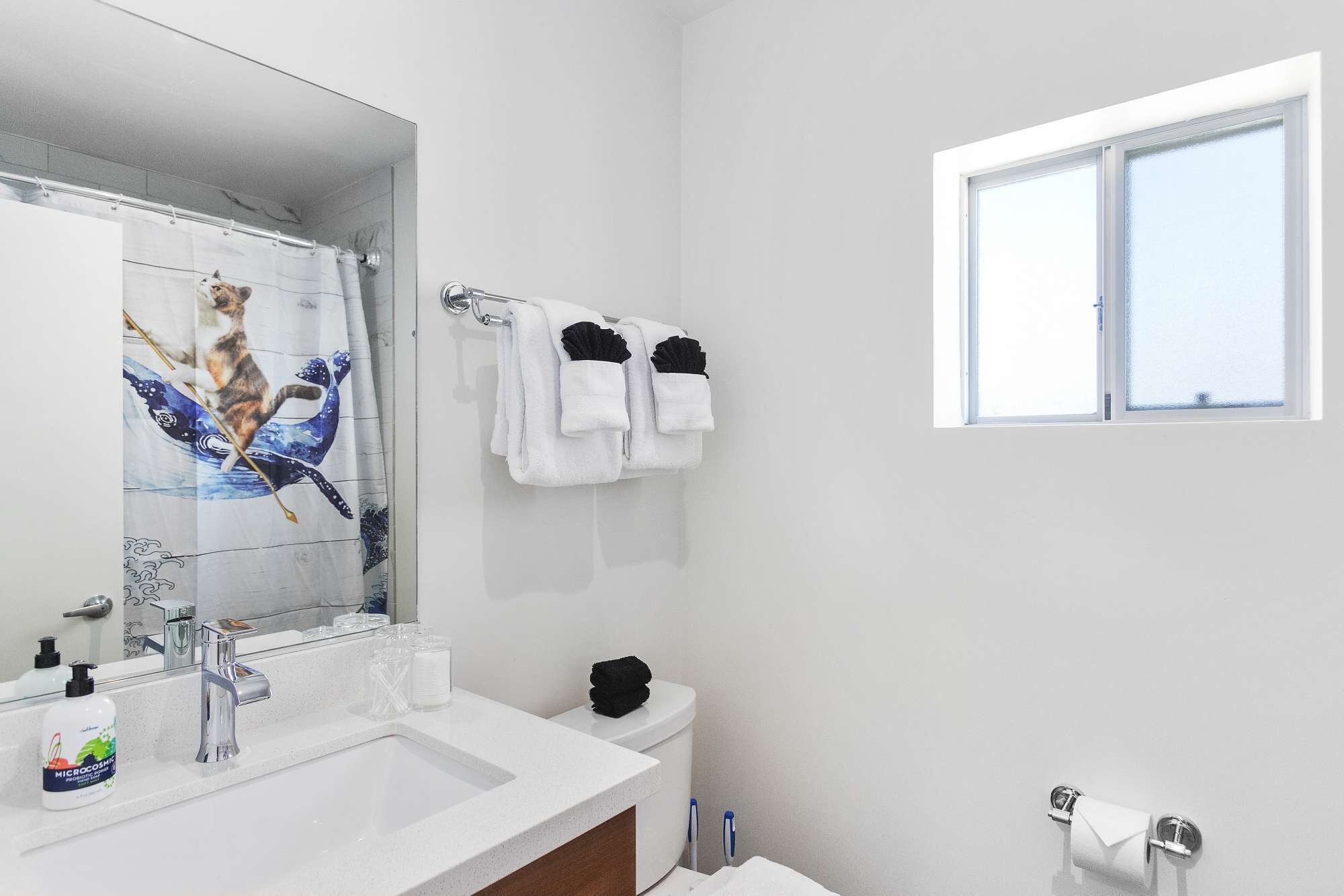 A modern bathroom with a large mirror, white sink, and a toilet. There is a window providing natural light and a towel rack with white towels.