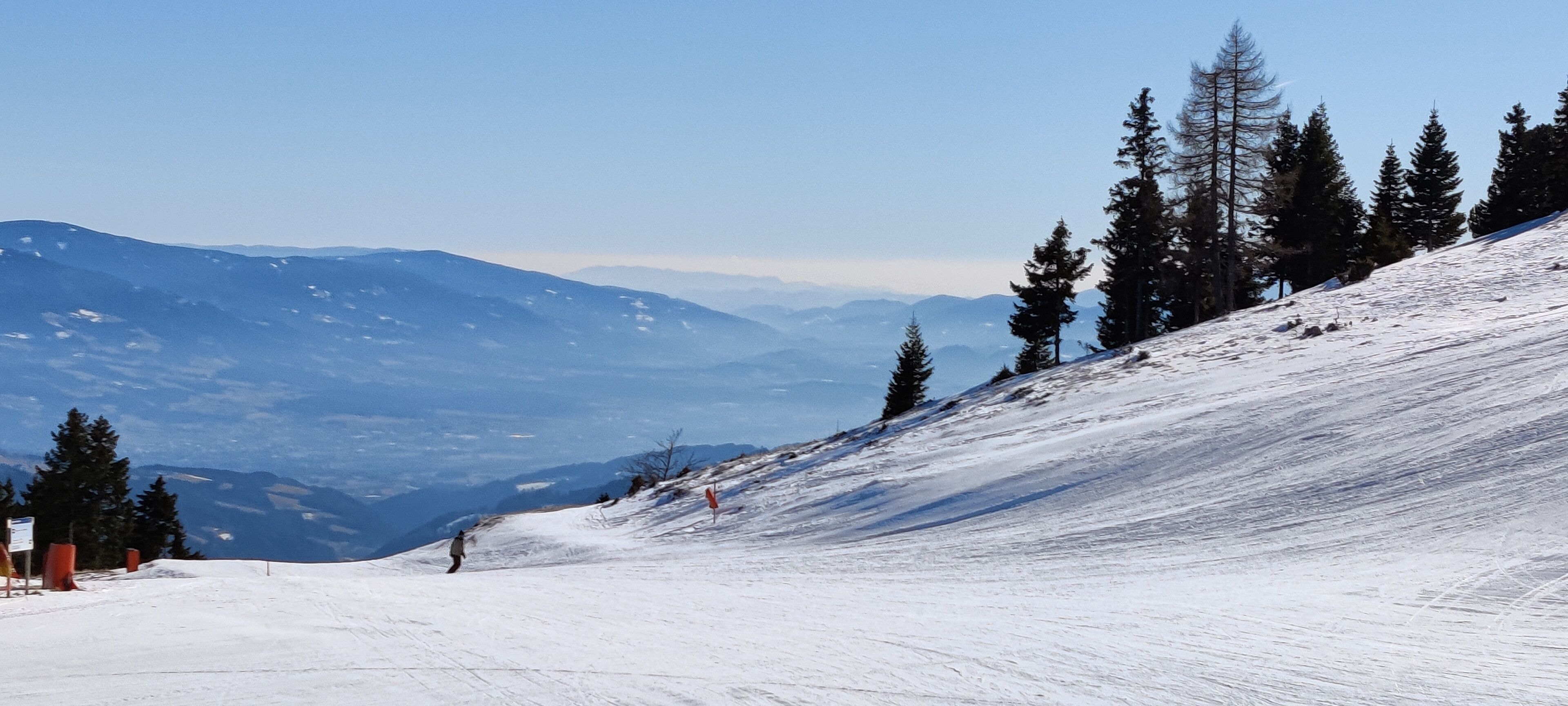A snow-covered ski slope with a clear view of distant mountains and evergreen trees.
