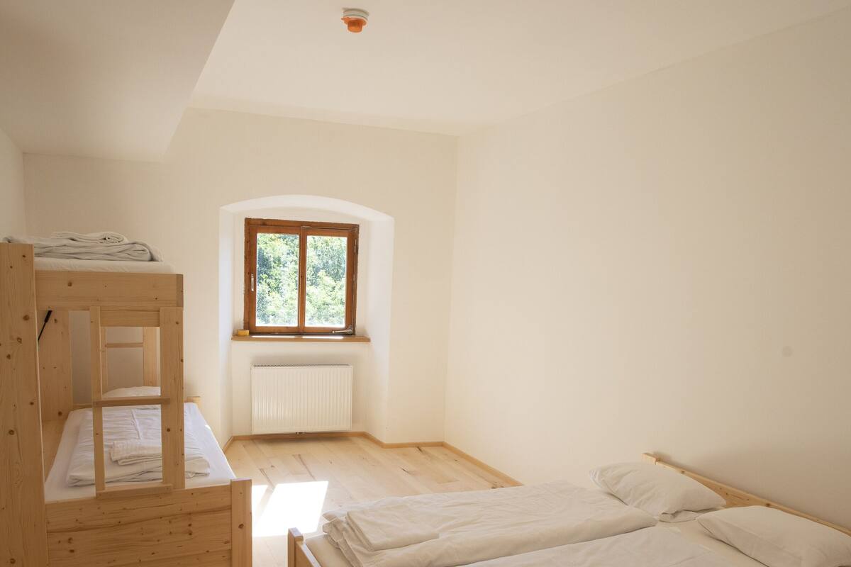 A small, bright room with a bunk bed and a single bed, a window with a view of greenery, and a ceiling light.