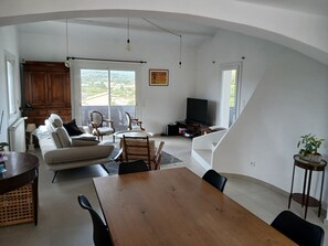 Uma sala de estar ampla com sofá, cadeiras, mesa de jantar, televisão e vista através de grandes janelas.