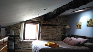 Een slaapkamer met een stenen muur, een bed met een bedovertrek met patroon, een raam met gordijnen en een houten dressoir.