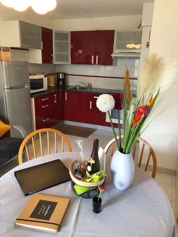 A modern kitchen with red cabinets, a microwave, and a sink. There's a dining table with a laptop, a book, and a bottle of wine. - Cosy apartment 100 m from Pentrez beach (Saint-Nic)