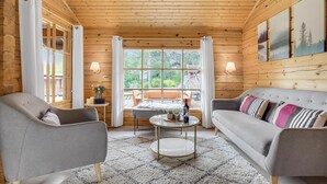 Indbydende interiør i en træhytte med træloft, to grå lænestole, en grå sofa med mønstrede puder, et rundt sofabord og et stort vindue med gardiner.