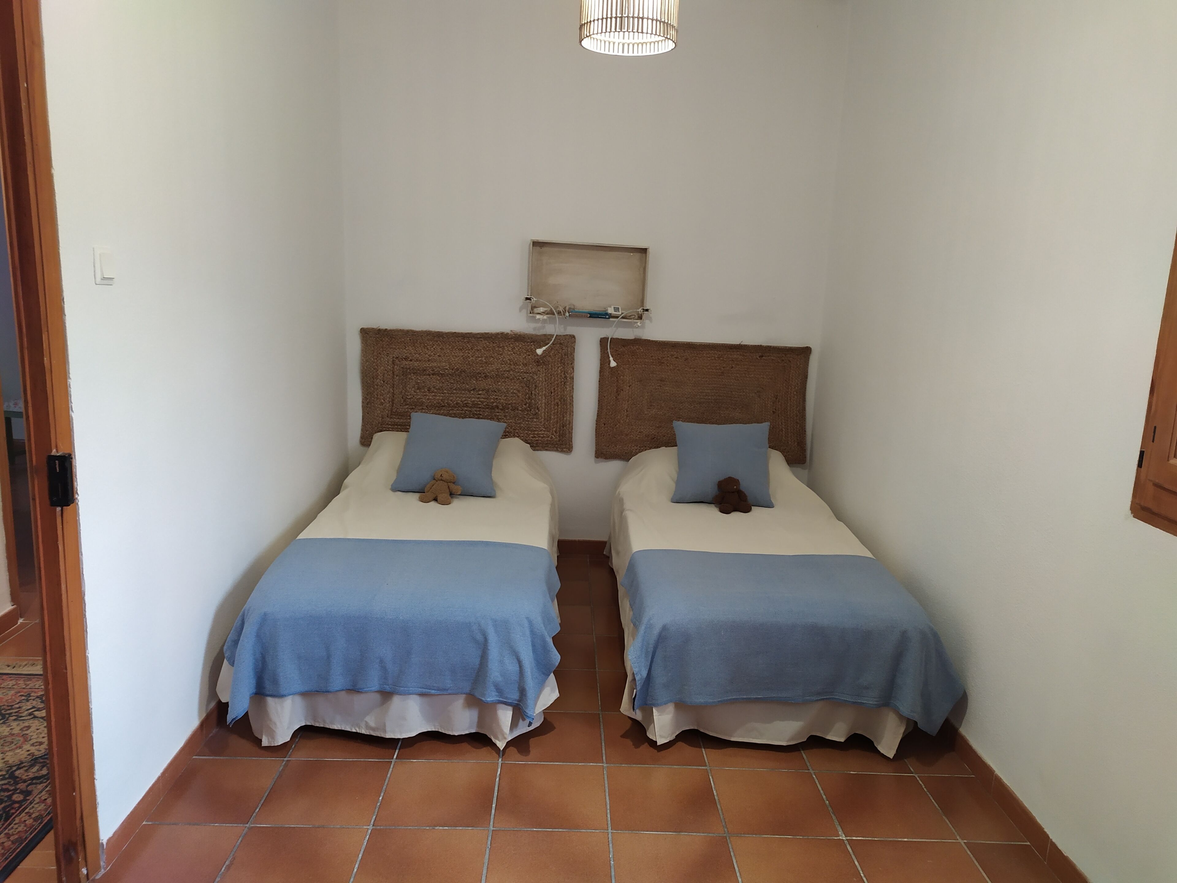 A small, clean room with two beds, each with a blue bedspread and pillows, and a woven headboard.