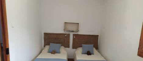 A small, clean room with two beds, each with a blue bedspread and pillows, and a woven headboard.