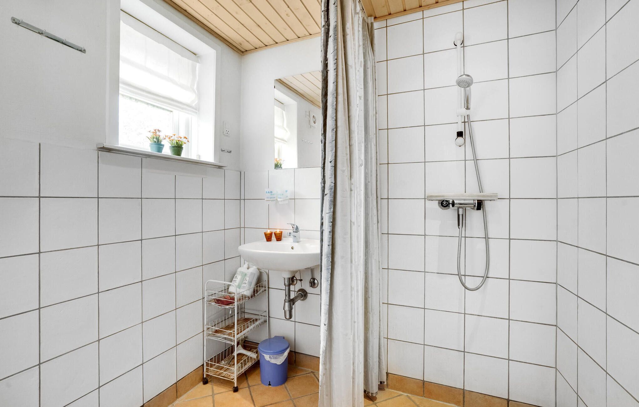 Un baño con paredes de azulejos blancos, una ventana con plantas, un lavabo, una ducha y un mueble de almacenamiento pequeño con un cubo de basura.
