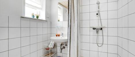 Un baño con paredes de azulejos blancos, una ventana con plantas, un lavabo, una ducha y un mueble de almacenamiento pequeño con un cubo de basura.