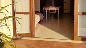 Una habitación con una cama, una mesa y sillas. Hay una ventana con cortinas, y una planta es visible en primer plano.