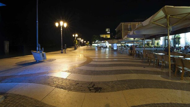 Un espacio al aire libre de noche con un sendero pavimentado, farolas y mesas al aire libre para un restaurante.