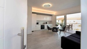 Eine moderne Küche mit weißen Schränken, ein Essbereich mit rundem Tisch und Stühlen sowie ein Ausblick ins Freie durch große Fenster.