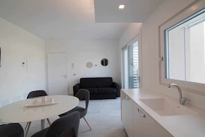 A modern kitchen with a white countertop and sink, a dining area with a round table and chairs, and a dark-colored sofa in the background.