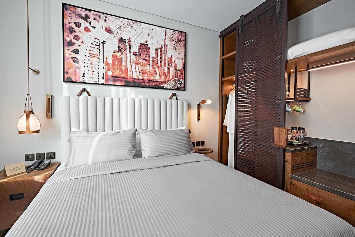 A modern hotel room with a large bed, a cityscape artwork, a wooden headboard, a nightstand with a phone, and a wooden side table with a mirror and a small shelf.