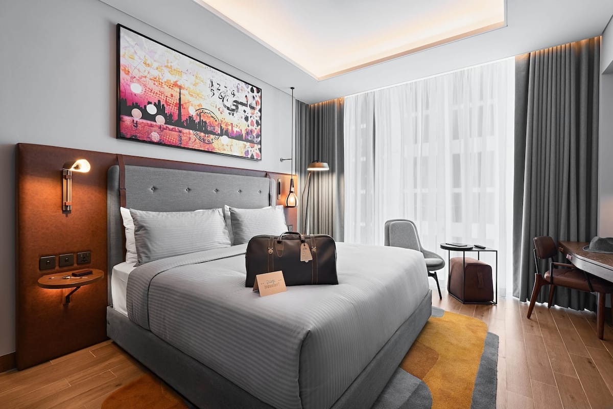 A modern hotel room with a large bed, a desk, and a chair. There is a painting on the wall, a lamp, and a suitcase on the bed.