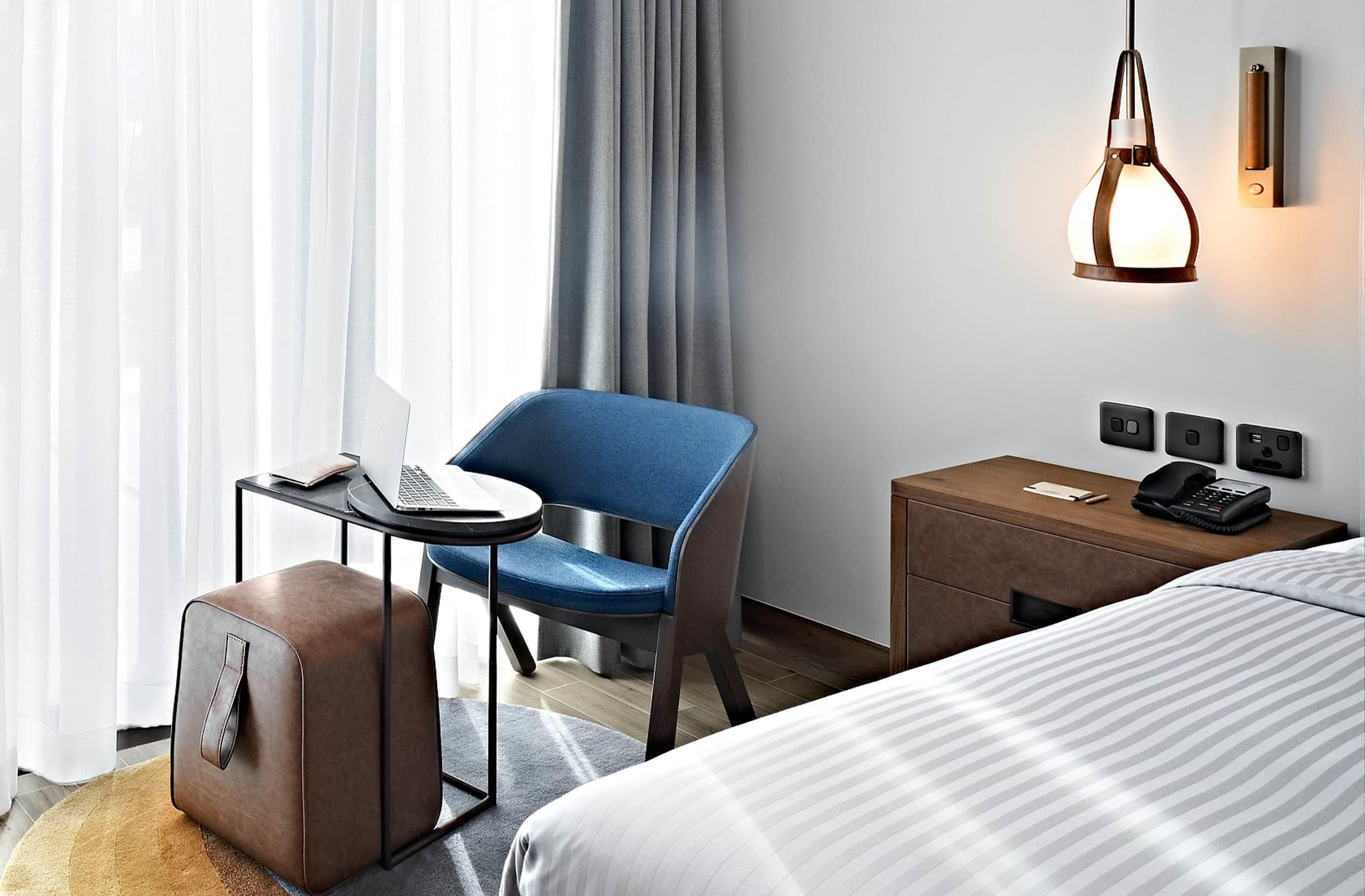 Ein Hotelzimmer mit einem Bett, einem Schreibtisch mit einem Laptop, einem Sessel, einem Koffer, einem Nachttisch mit einem Telefon und einer Wandleuchte.