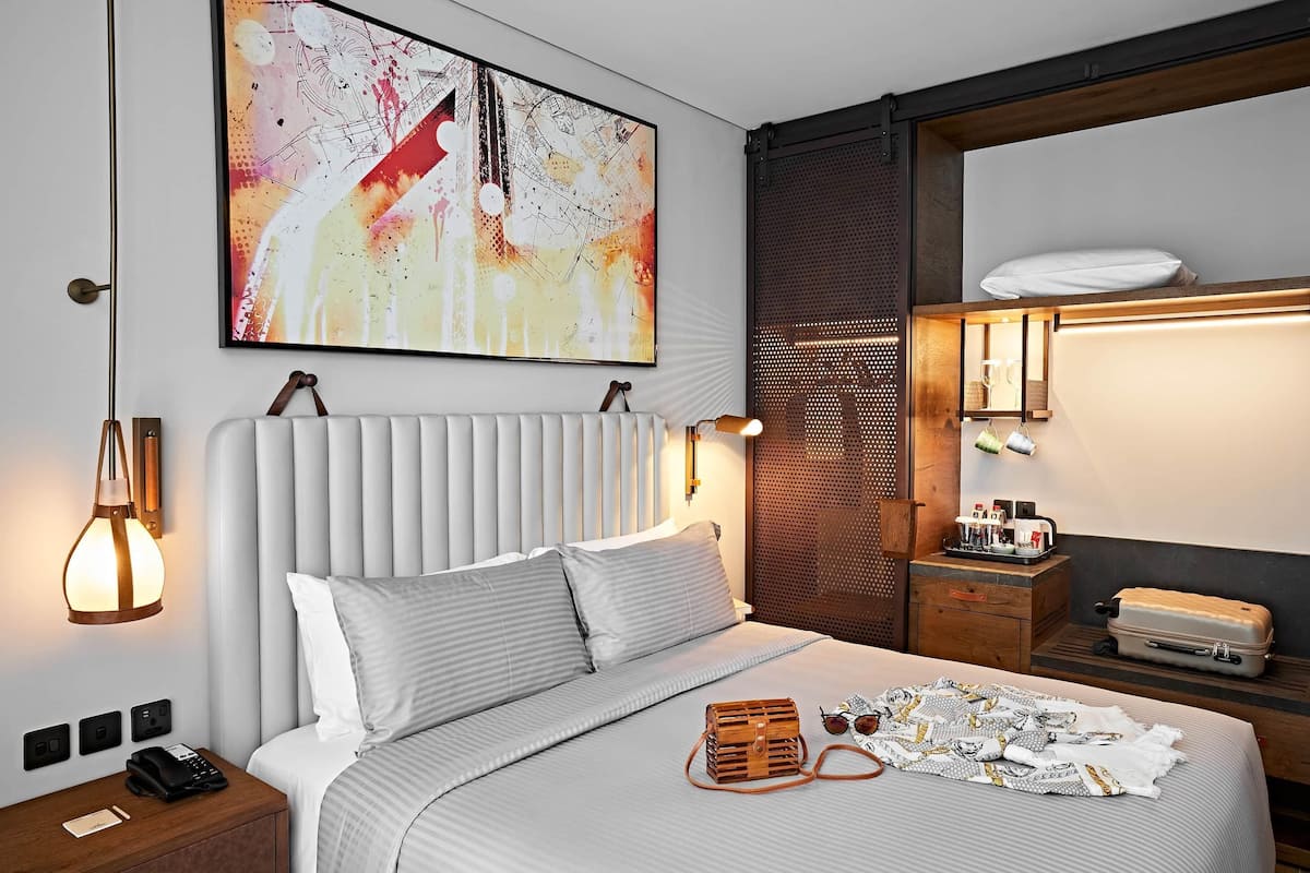 A modern hotel room with a large bed, a bedside table with a lamp, a wall-mounted artwork, and a suitcase on the floor.