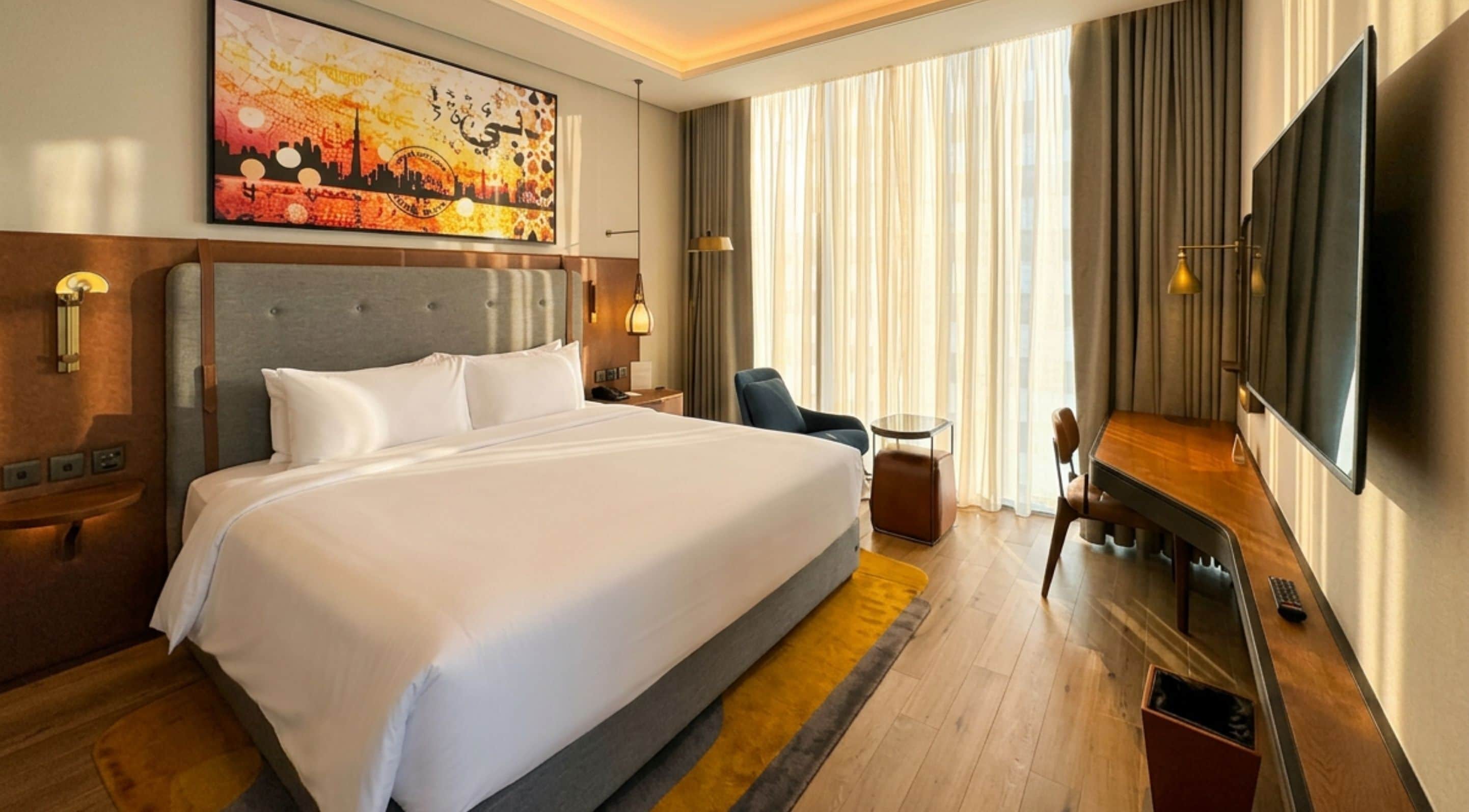 A hotel room with a large bed, a desk with a chair, a television, and a painting on the wall.