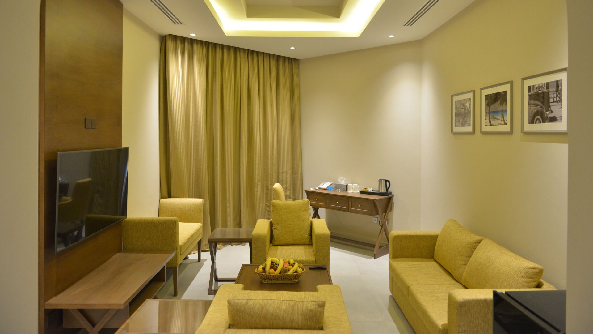 A modern living room with a sofa, armchairs, a coffee table with fruit, a desk, and a television.