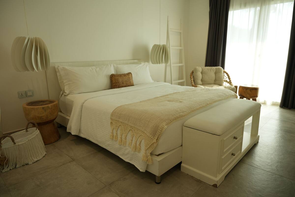Un dormitorio con una cama, mesitas de noche, una silla, un taburete de madera y un bolso tejido.