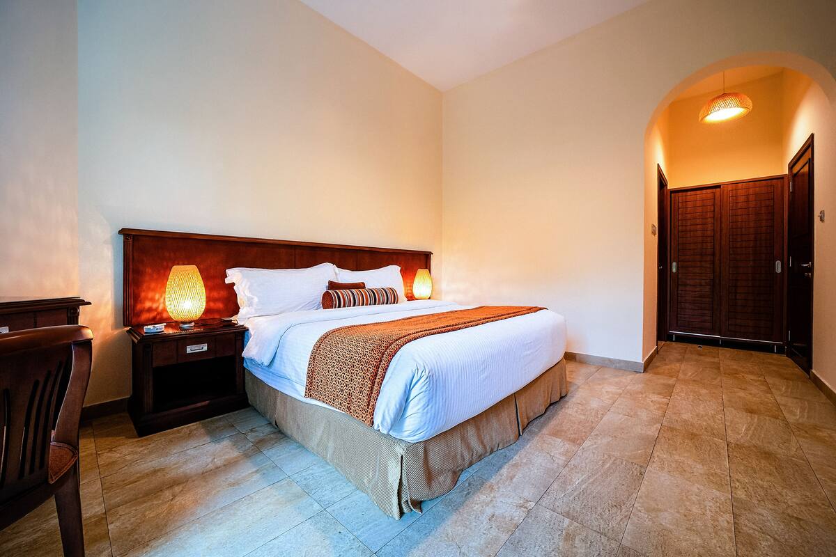 Una habitación de hotel con una cama grande, cabecera de madera, dos lámparas de noche, una mesita de noche y un armario.