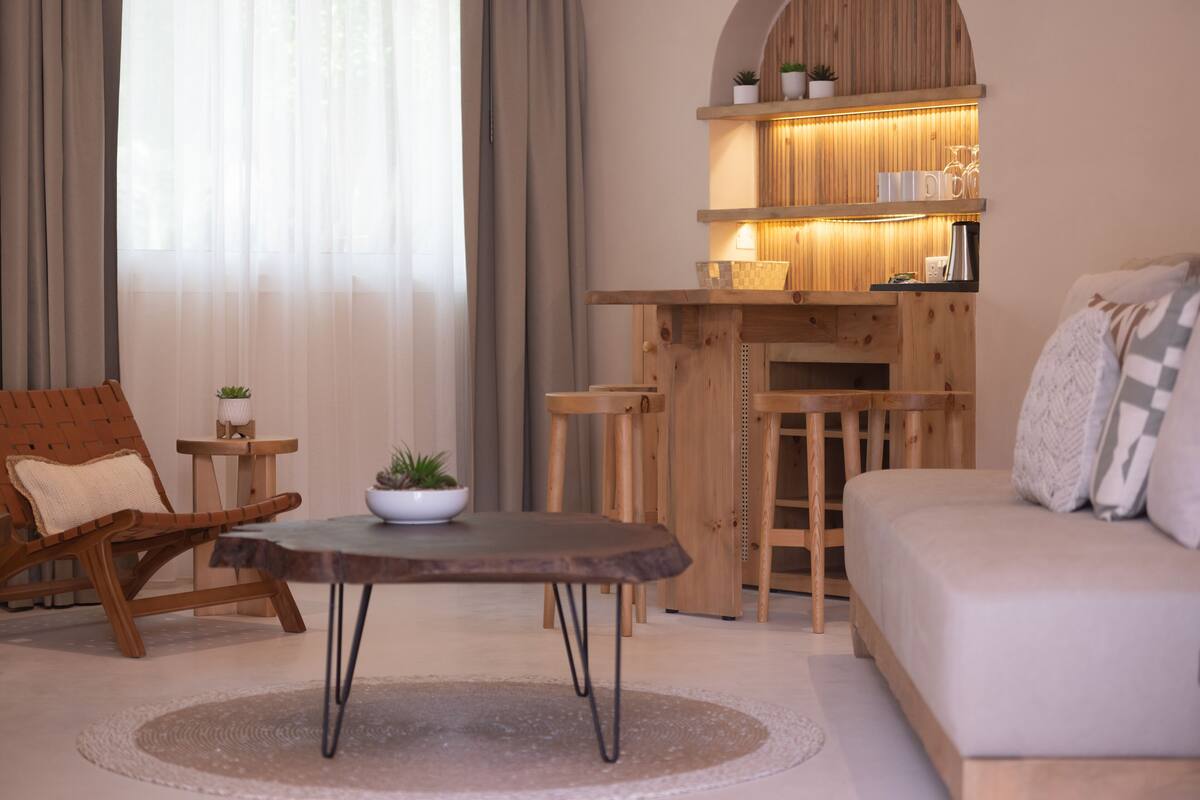 Una sala de estar acogedora con un sofá de madera, una mesa redonda y una estantería de madera con plantas y objetos decorativos.