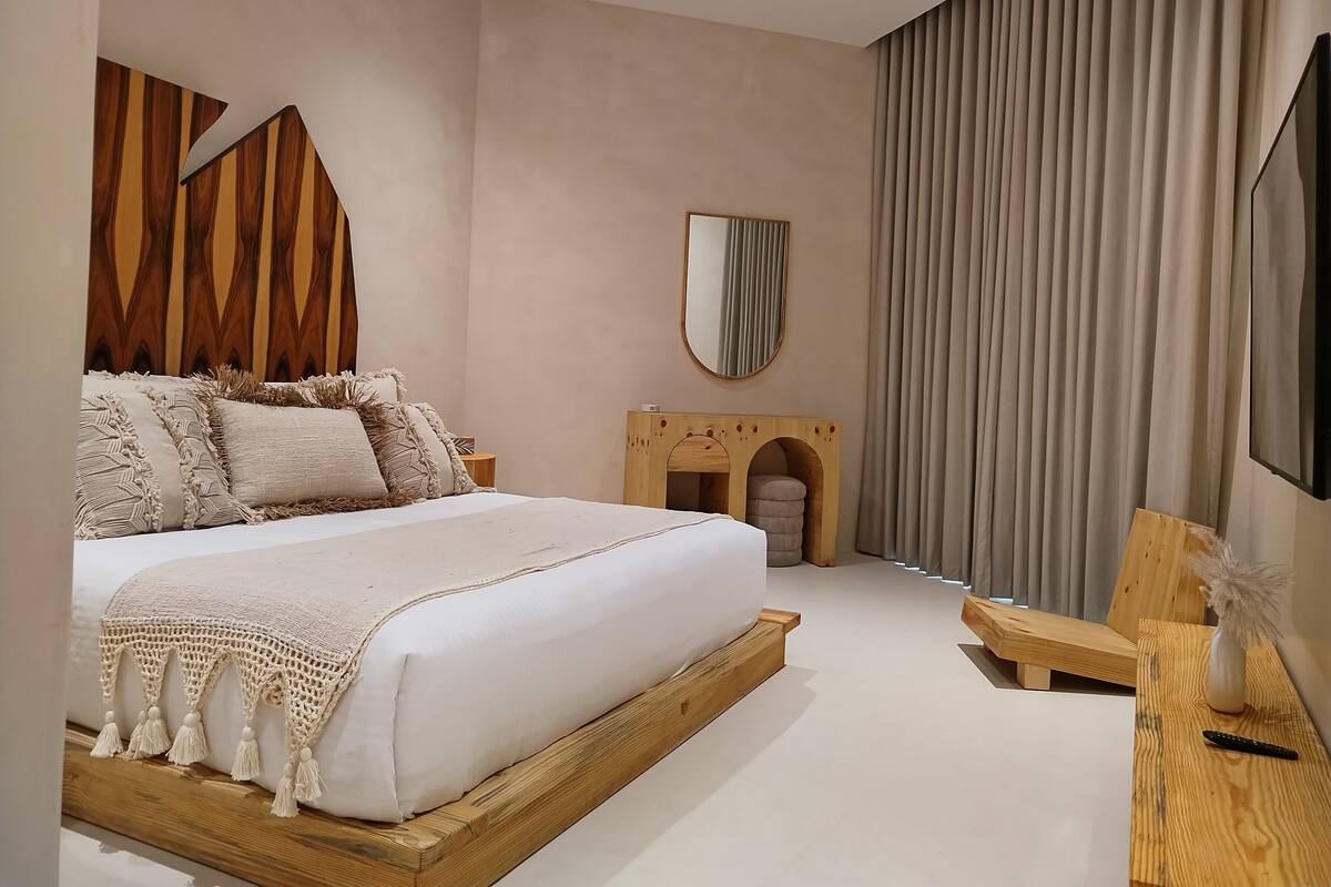 Un dormitorio moderno con una cama de madera, un pequeño armario de madera, un espejo y un televisor sobre un mueble de madera.