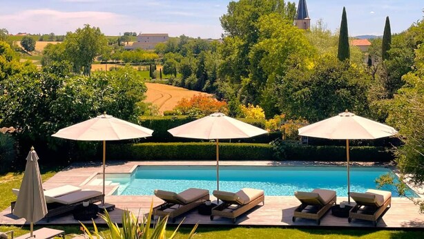 A pool with lounge chairs and umbrellas, surrounded by greenery and a distant church. - Le Domaine de la Monestarié<br>Charm, elegance and scents of Tuscany (BERNAC)