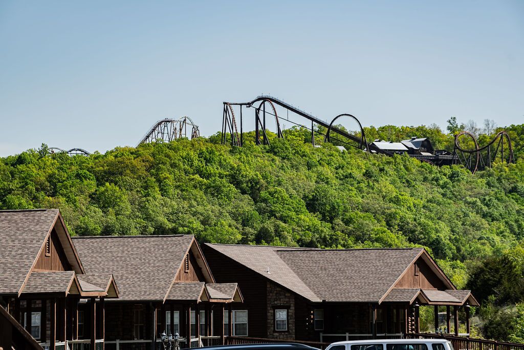 A rustic lodge with multiple wooden cabins, a roller coaster in the background, and a lush green hillside.