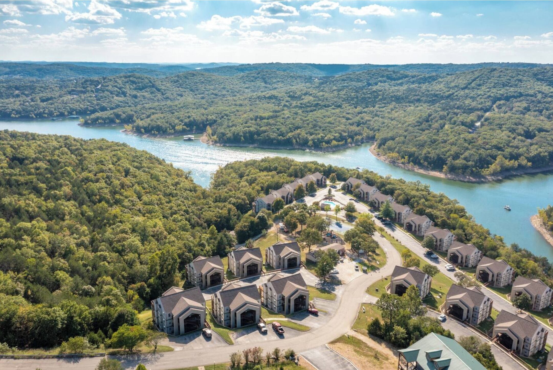 Vue aérienne d’un quartier résidentiel avec de nombreuses maisons à deux étages, une route sinueuse et un lac entouré d’une forêt dense.