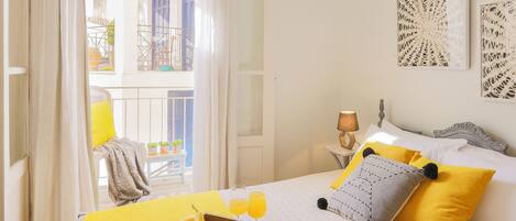 Una habitación de hotel luminosa con una cama, almohadillas amarillas, una bandeja con plátanos y vasos de zumo de naranja, y un balcón con contraventanas azules.