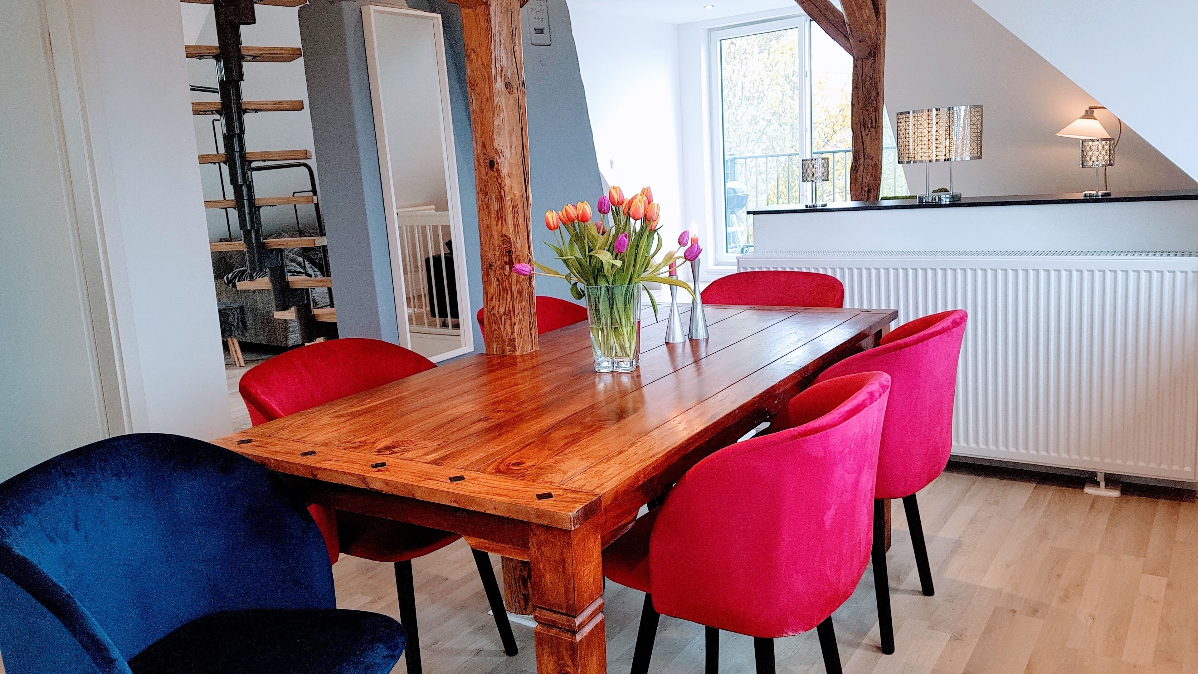 Ein Essbereich mit einem Holztisch, roten und blauen Stühlen sowie einer Blumenvase.