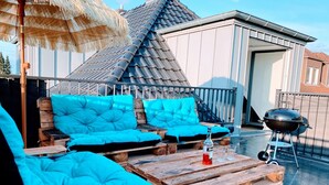 Une terrasse sur le toit avec un banc en bois et des coussins, un barbecue et un parasol en chaume.