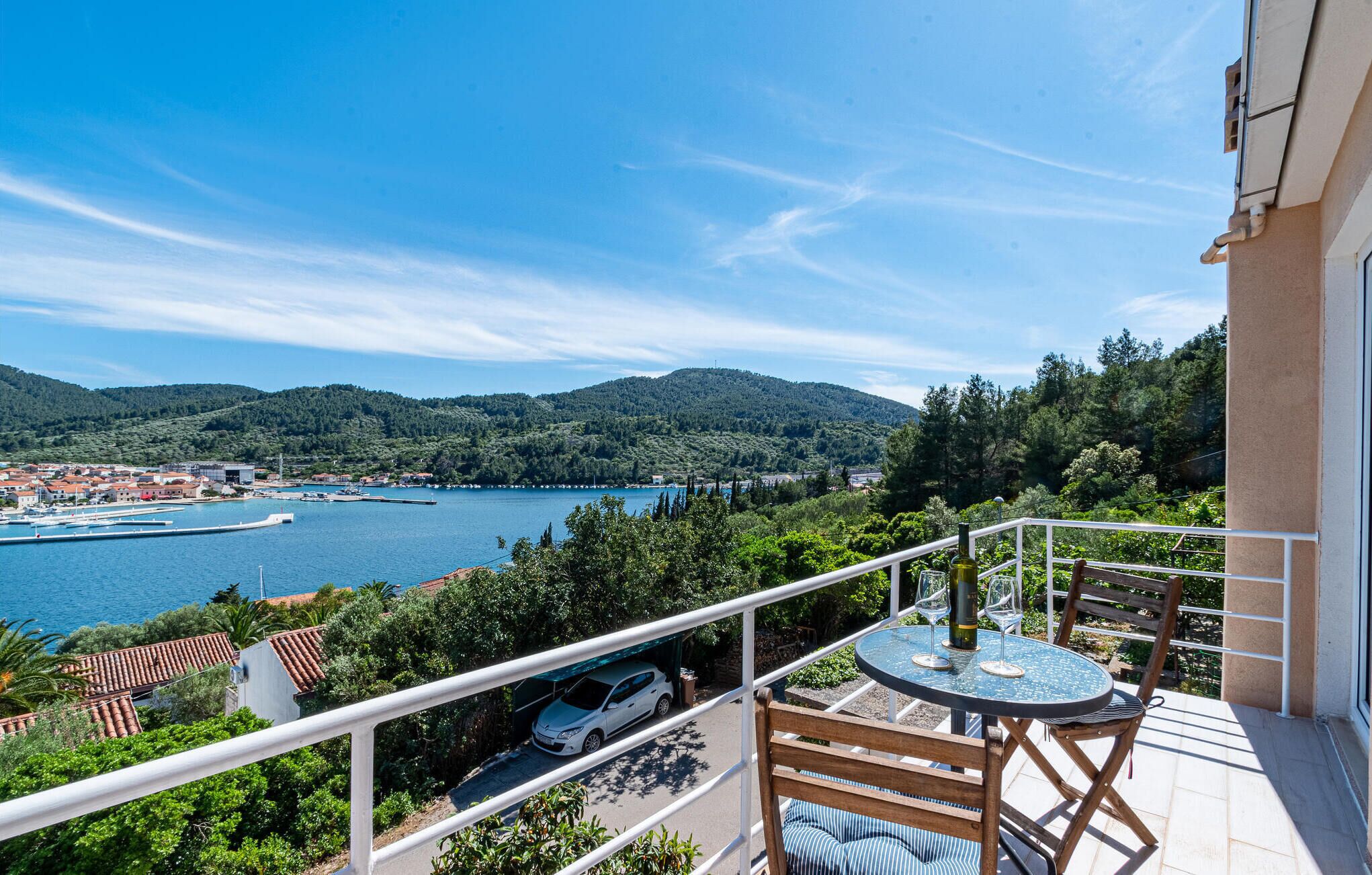 A balcony with a table and chairs overlooking a lake, surrounded by greenery and buildings.