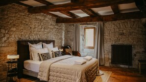 A bedroom with a stone wall, a bed with multiple pillows, a bedside table with a lamp, and a window with curtains. - Le Domaine de la Monestarié<br>Charm, elegance and scents of Tuscany (BERNAC, TARN)