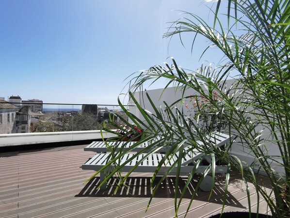 A rooftop terrace with sun loungers, a potted plant, and a view of the cityscape.