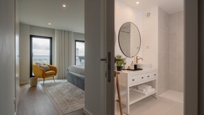 Une chambre d’hôtel moderne avec un grand lit, un fauteuil jaune, une coiffeuse blanche avec un miroir et une vue sur la mer par la fenêtre.