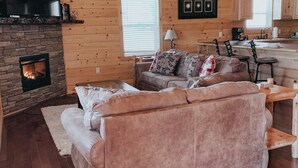 A cozy cabin interior with a fireplace, wooden walls, a sofa, and a dining area. - Twin Oaks: Close to Town! Mtn Views! Hot Tub!  (Bryson City)