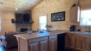 A wooden cabin interior with a kitchen island, built-in cabinets, a stone fireplace, and a large window. - Twin Oaks: Close to Town! Mtn Views! Hot Tub!  (Bryson City)