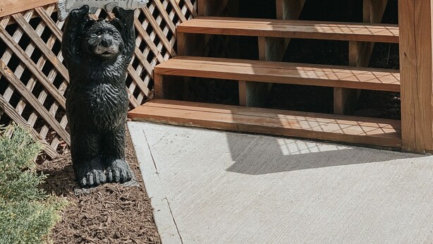 A wooden staircase leading to a covered area with a metal fence and a bear statue with a fish on its head. - Twin Oaks: Close to Town! Mtn Views! Hot Tub!  (Bryson City)