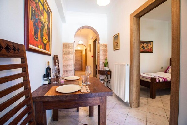A dining area with a wooden table set for two, a chair, and a bottle of wine. There is a painting on the wall and a door leading to another room.