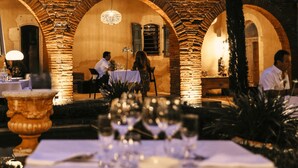 An outdoor dining area with brick arches, tables set with white tablecloths and wine glasses, and a chandelier hanging from the ceiling. - Le Domaine de la Monestarié<br>Charm, elegance and scents of Tuscany (BERNAC, TARN)